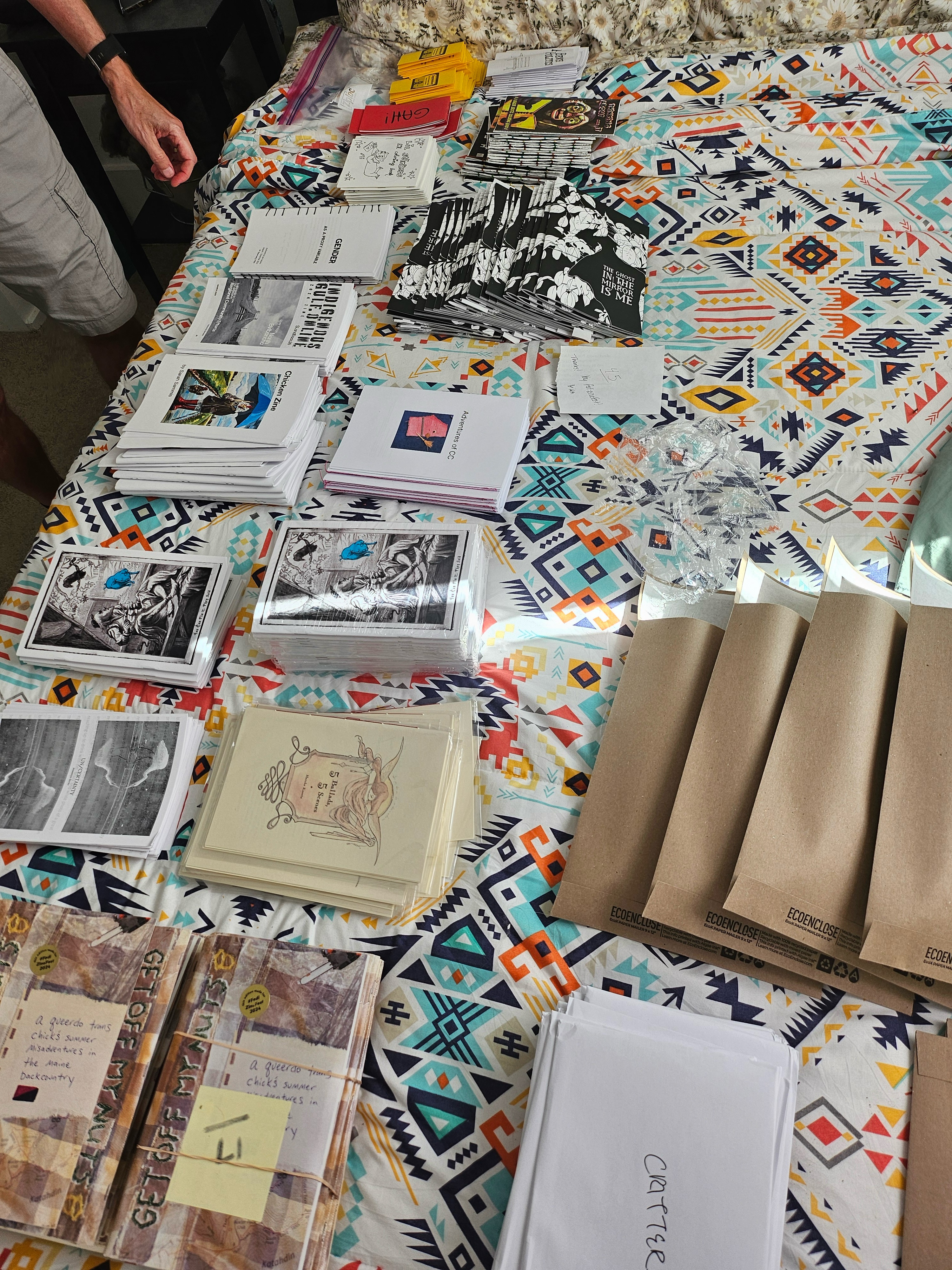 A number of zines of various sizes and designs and colors laid out on a very colorful bedspread.