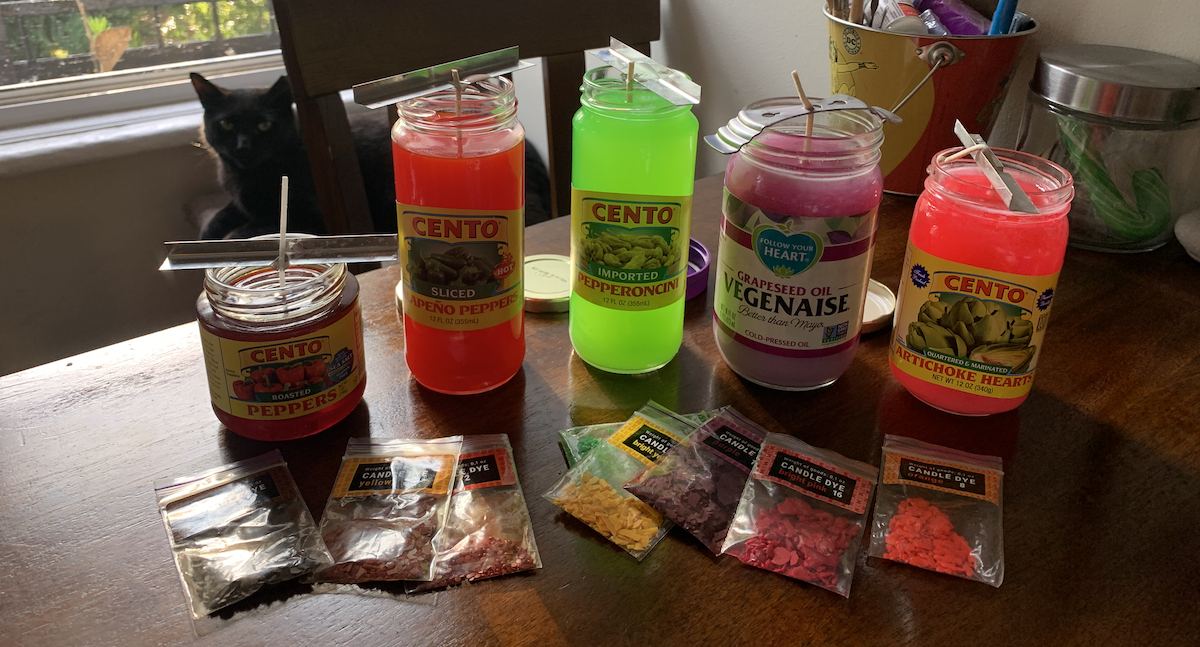 a photo of my kitchen table, with my black cat pumagreg in the background, covered in jar lids, wax dye flakes in little bags, and newly-poured colorful candles that are still hot so the colors are very neon and vibrant