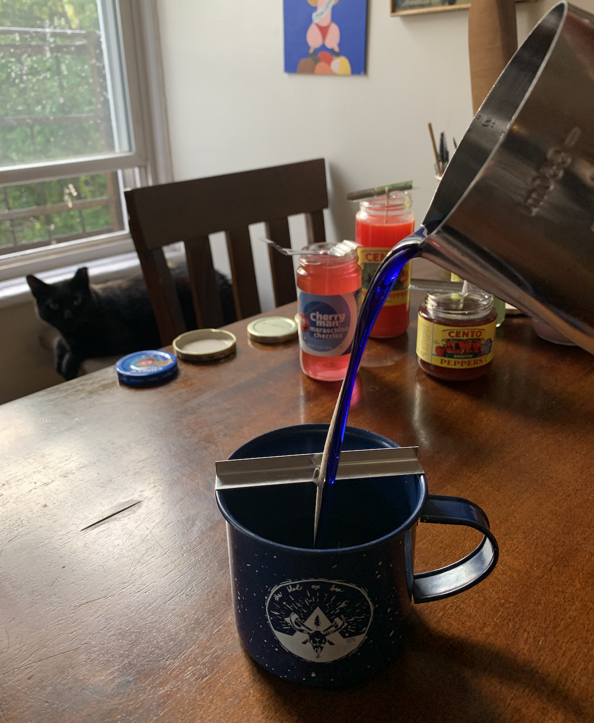 a photo of my kitchen table, with my black cat pumagreg in the background, and i'm in the middle of pouring a dark blue wax into a matching blue tin mug