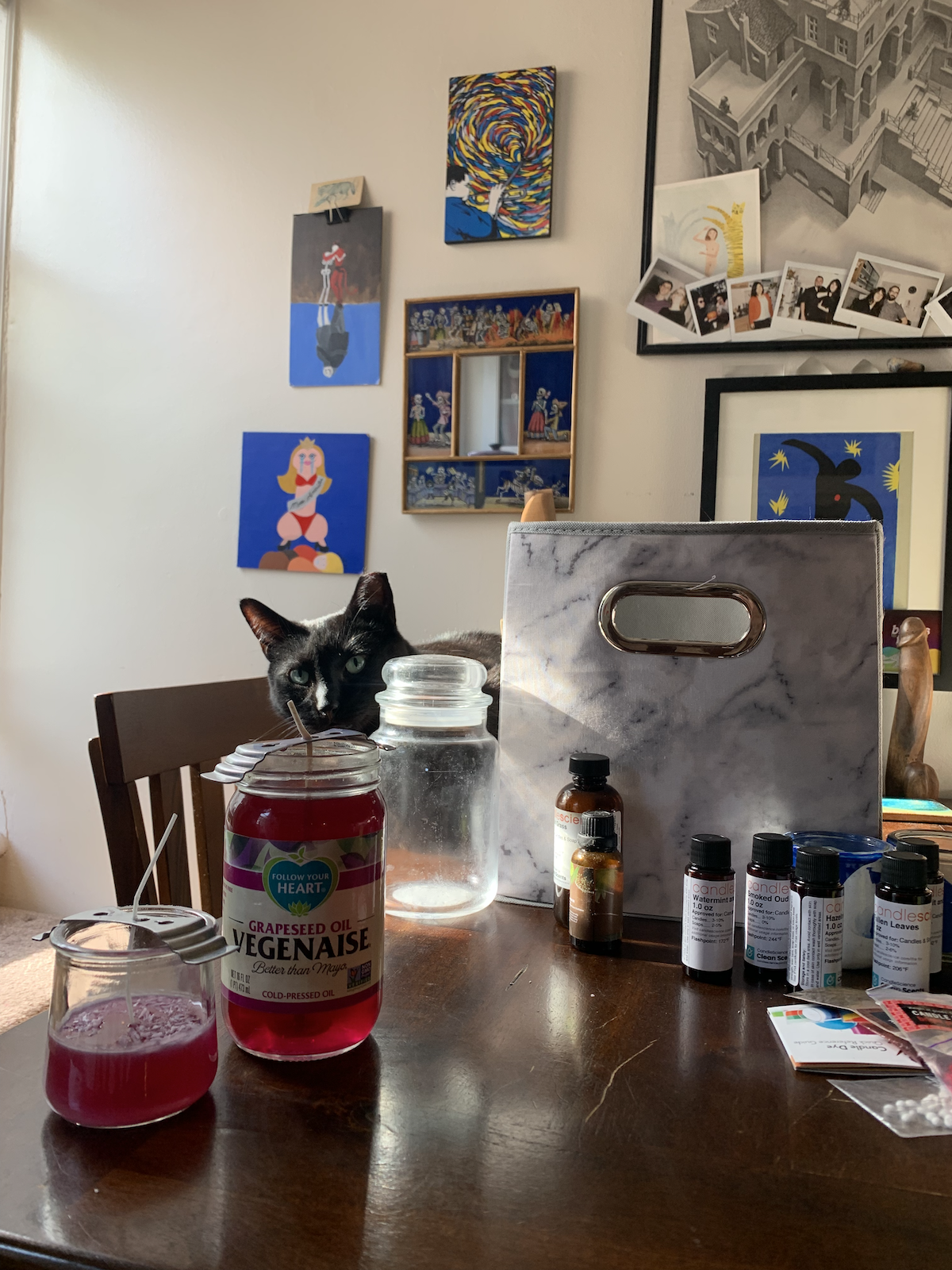 a photo of my kitchen table, with my black cat pumagreg in the background, with a veganaise jar full of hot purple wax and there are a bunch of small fragrance oil bottles and wax dye flakes in little bags