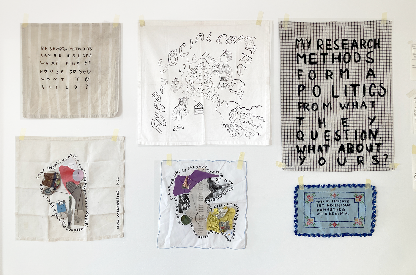 Six pieces of fabric using mixed media collage, embroidery and black ink. Six pieces of fabric are shown hanging on a white wall. From top left, the first piece is a grey fabric
with white stripes, with the following sentence embroidered 'Research methods can be brics. What kind of
house do you want to build?'. The second piece, on the top middle row is a white cotton napkin that has
been painted with black ink. It reads in wavy bulky outlined letters'Food as a social construct' and shows
four figures holding and extending food. One of these figures is annotated with a speech bubble coming from
their open mouth that reads 'Hunger is also gendered'. This same figure's wavy hair is annotated with the words
saciation, hedonics, homeostasis, and cuando cuando cuando. The third pice on the top row, on the right far corner is
a black and white squared patterned kitchen cloth with the sentence 'my research methods form a politics from whaty
they questions. What about yours?' painted with black in. On the bottom row, the first piece on the left shows a
mixed media collage on a white kitche cloth and is annotated by the following sentence surrounding the collage:
'A sentence that encapsulates a story larger than itself. Mariana Pestana.' Next to it,in the middle bottom row,
there's another mixed media collage with the sentence: 'A space where all your needs are met. Hand holding, dancing,
laughing, eating, doing, being, together. Mas interdependencia.' The final piece, bottom-left, is a rectangular light
blue little table cloth, with blue embroidery trimmings, and flowers. The sentence 'Viver um presente sem necessidade
dum futuro que o redima' (translation from portuguese: To live a present without the need of a future to redeeem it)
in black ink.